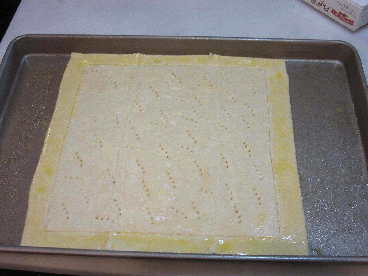 A square of puff pastry dough with an egg washed border and a docked center on a baking sheet before baking.