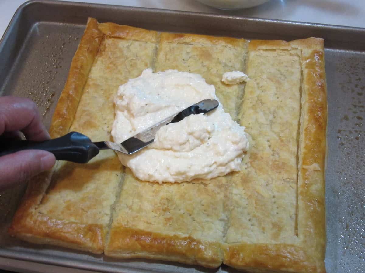 An offset spatula spreading a mayo, ranch, and Parmesan filling onto a sheet of baked puff pastry dough.