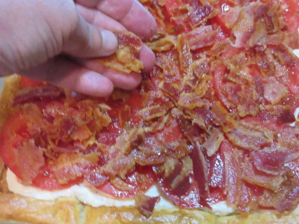 A hand spreading cooked, crumbled bacon over the top of seasoned tomato slices.