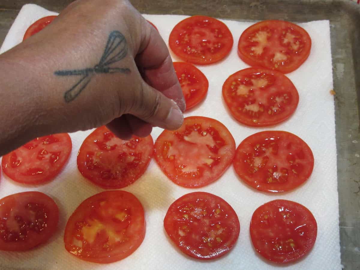 A hand adding salt to tomato slices lined up on a paper towel.