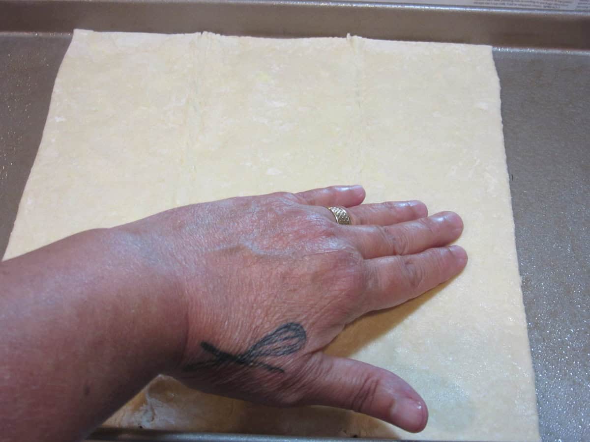 A hand pressing out a square of puff pastry dough.