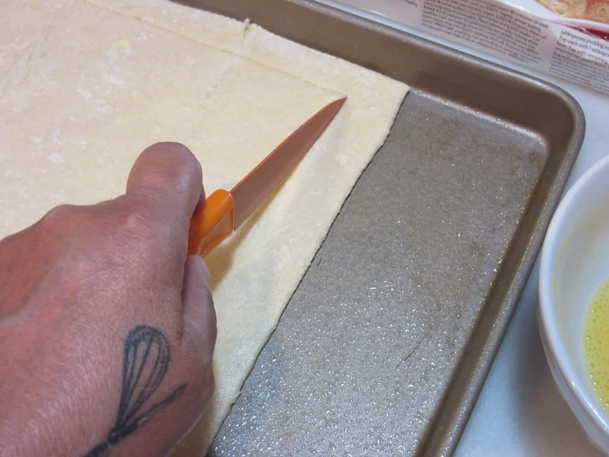A hand holding a paring knife cutting out a frame from a sheet of puff pastry dough.