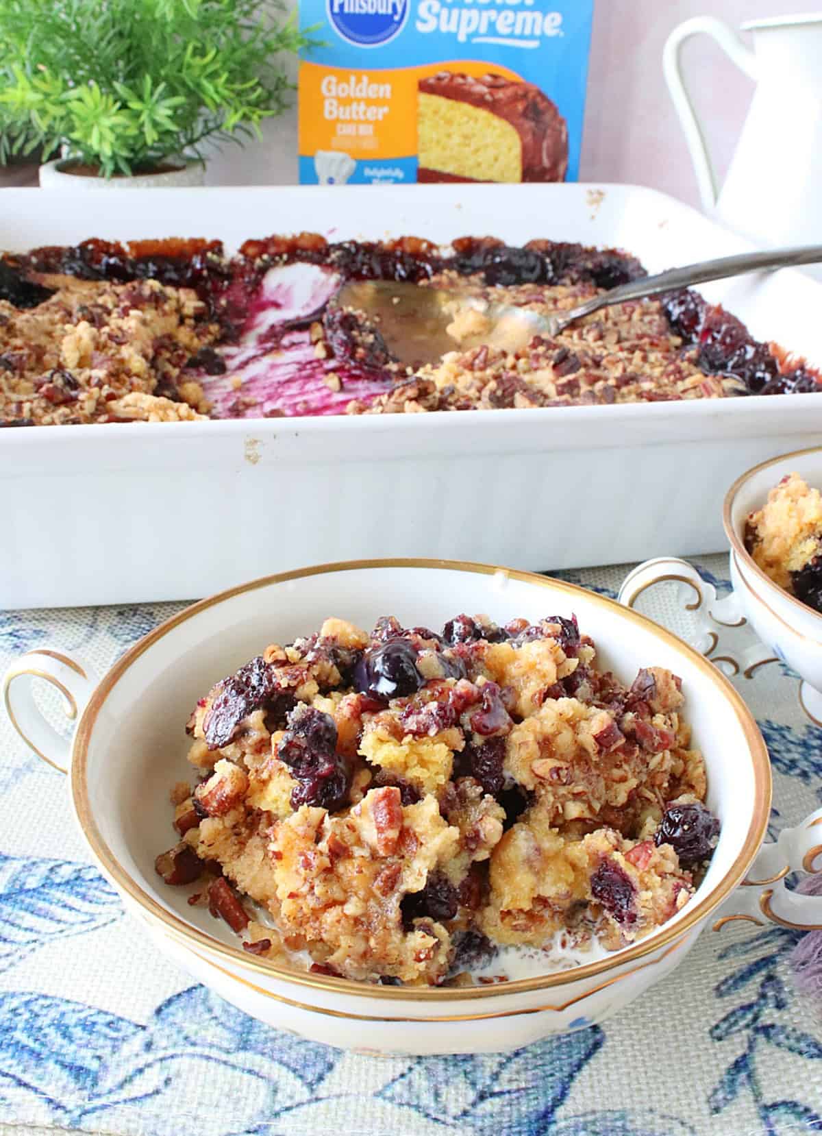 A serving of blueberry crisp in a small dish with the baking dish containing the remaining crisp in the background.