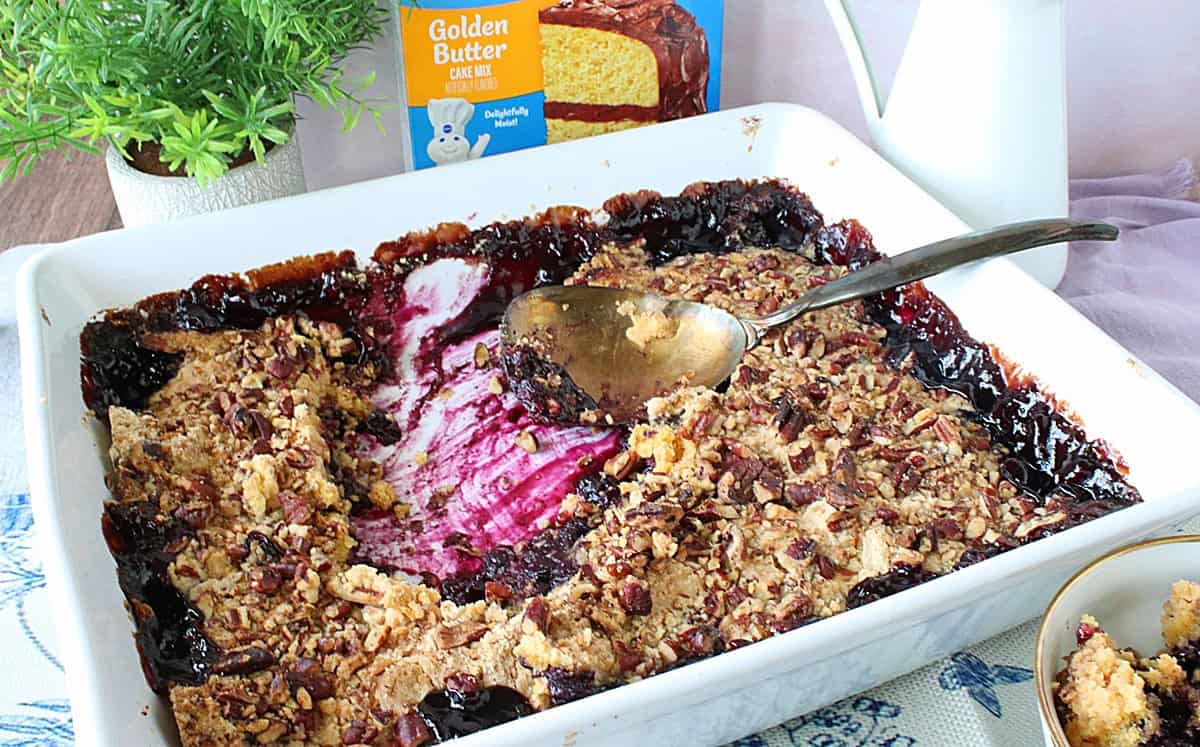 A freshly baked cake-mix blueberry crisp with bubbly blueberries and a crunchy topping on a table with a serving spoon.