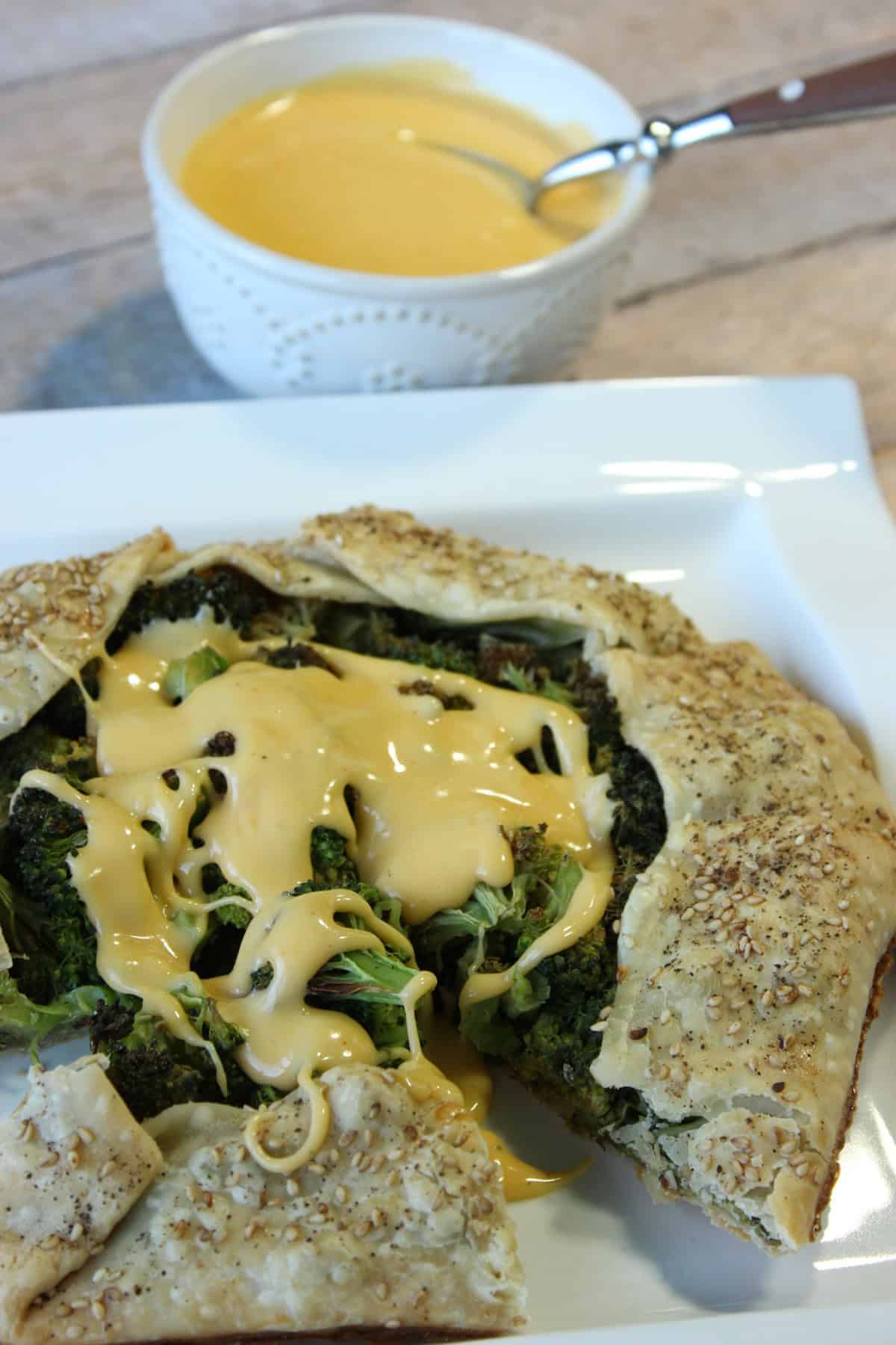A broccoli and cheese crostata on a plate with a bowl of cheese sauce in the background.