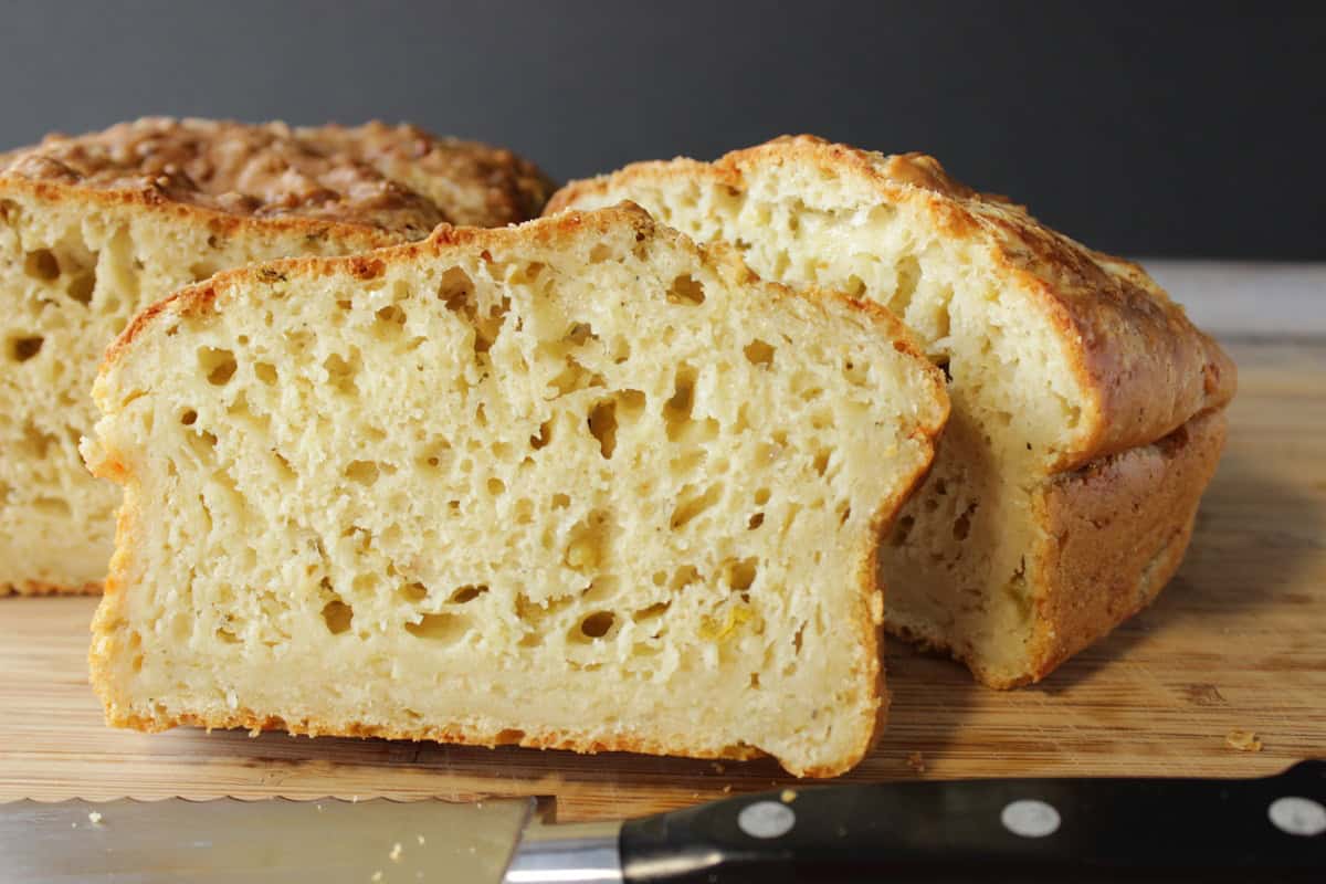 A serrated knife in front of a sliced loaf of cheese jalapeno quick bread on a wooden cutting board.