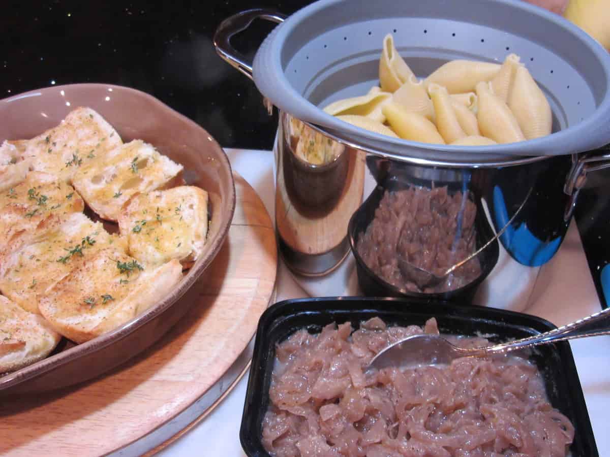 A casserole dish with bread, colander with pasta shells, and dish with red wine caramelized onions on a countertop.