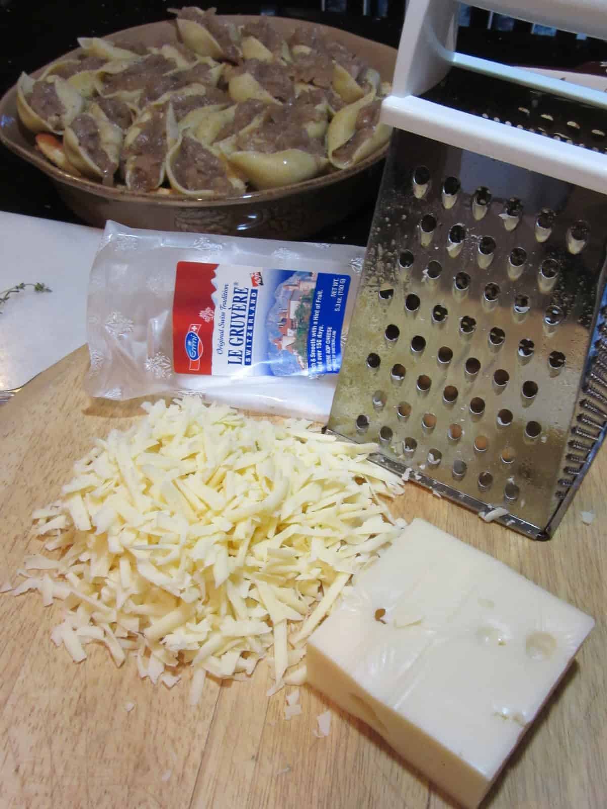 A box grater along with shredded Gruyere and Swiss cheese on a wooden cutting board.