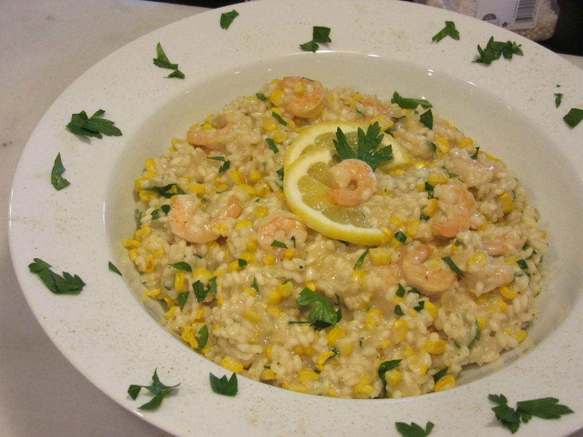 New England Sweet Corn Risotto is a great way to use up the last of summer's bounty of sweet corn. It's a creamy and delicious main course that can be on the dinner table in approximately 30 minutes. - Kudos Kitchen by Renee - kudoskitchenbyrenee.com A white bowl filled with shrimp and corn risotto with parsley around the side of the bowl.