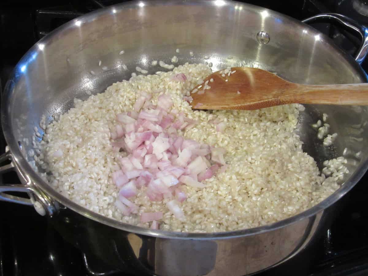 Arborio rice and chopped shallots in a large silver skillet with a wooden spatula.