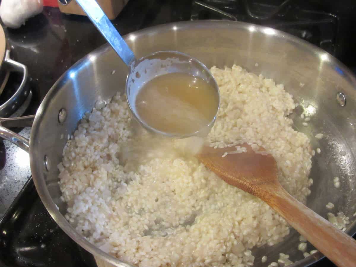 A ladle of hot chicken broth being added to a skillet of arborio rice.