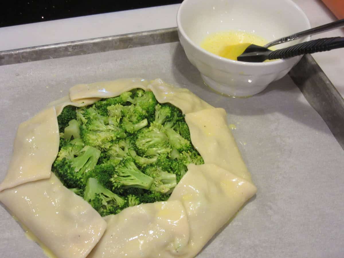 Pie crust after being egg washed surrounding cooked broccoli on a baking sheet.