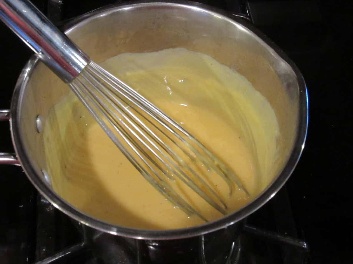 A whisk in a saucepan of creamy cheese sauce on the stovetop.