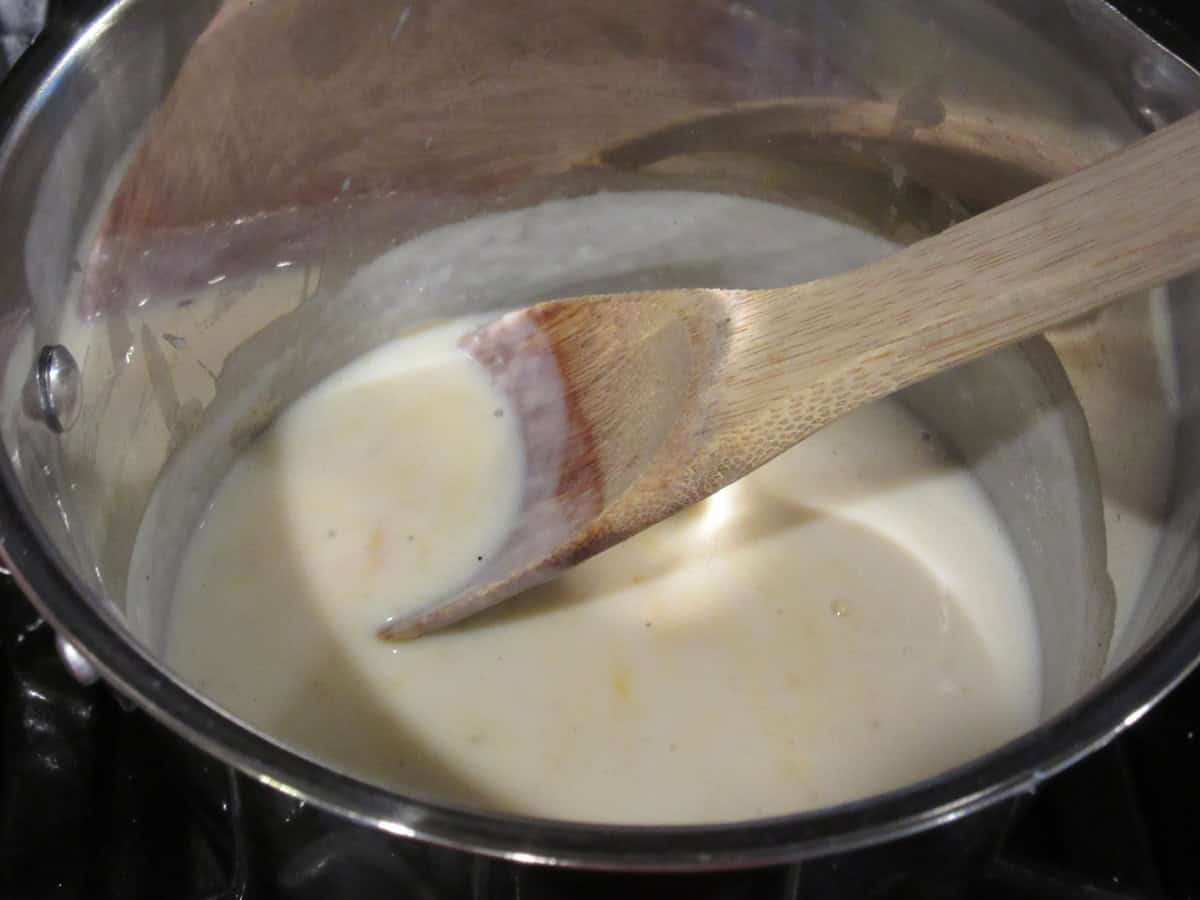 A wooden spoon with the beginnings of a cheese sauce in a saucepan.