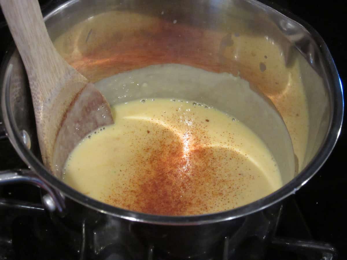 A wooden spoon in a saucepan with cheese sauce along with cayenne red pepper.