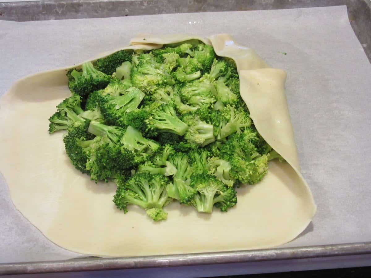Pie crust dough being folded around the edges of a bunch of cooked broccoli to make a savory crostata.