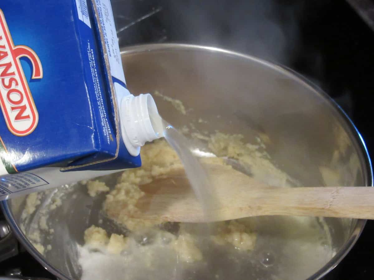 Chicken stock being poured into a saucepan with a thickened roux.