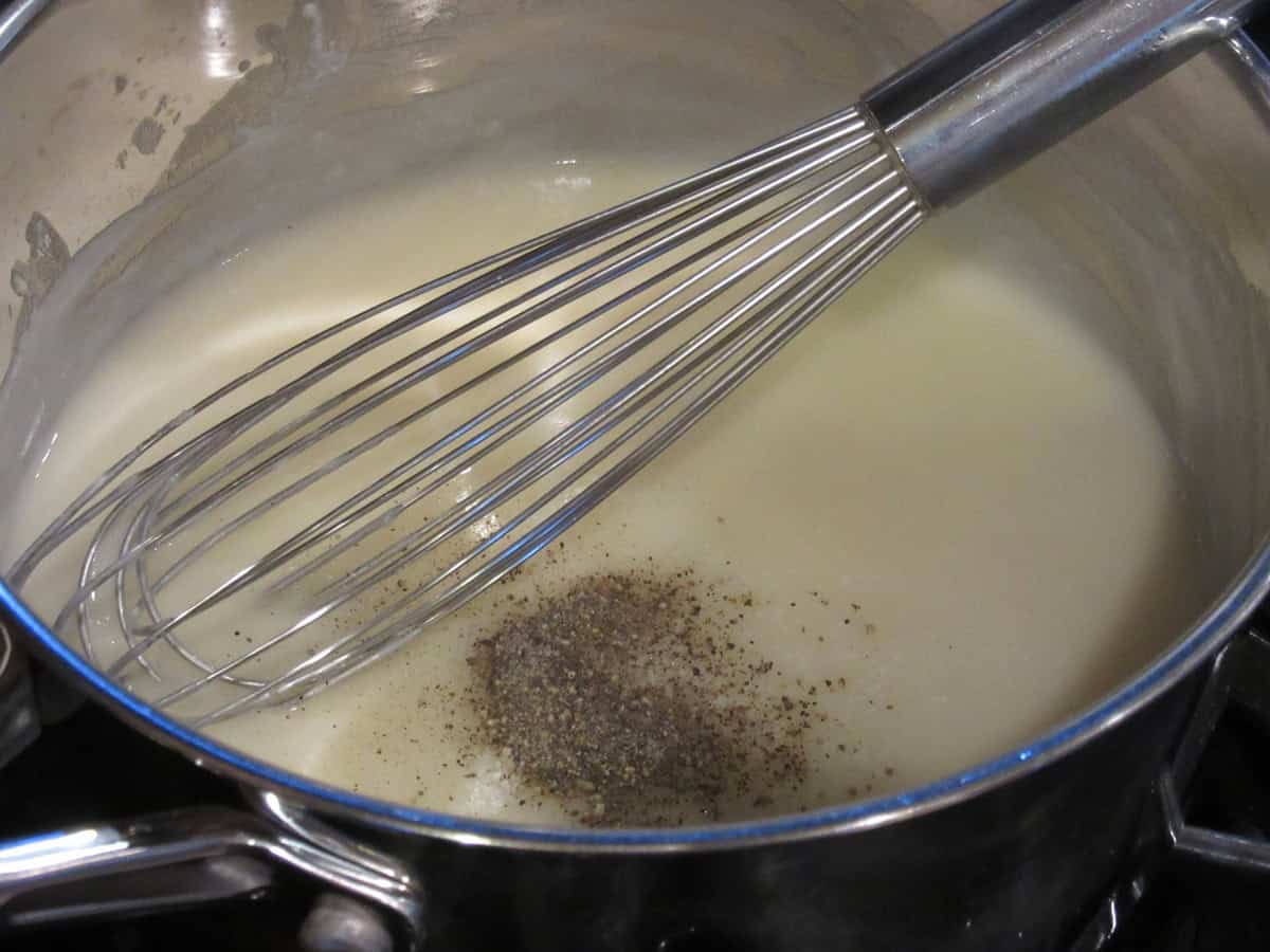 A thickened chicken base in a saucepan with black pepper and a whisk.