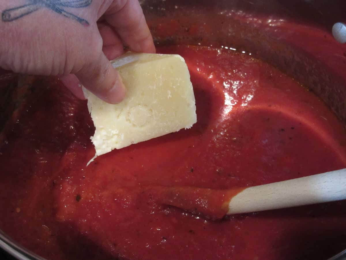 A rind of Parmesan cheese being added to a saucepan of homemade marinara sauce.