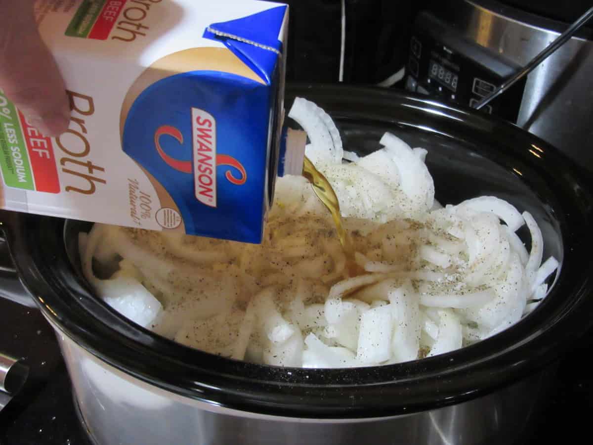 Beef broth being added to a slow cooker with sliced onions.