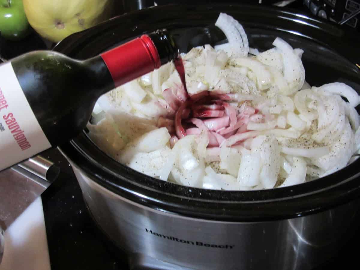 Red wine being poured into a low cooker filled with sliced onions.