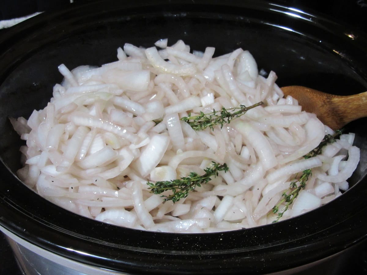Sliced onions and thyme sprigs in a slow cooker with a wooden spoon.