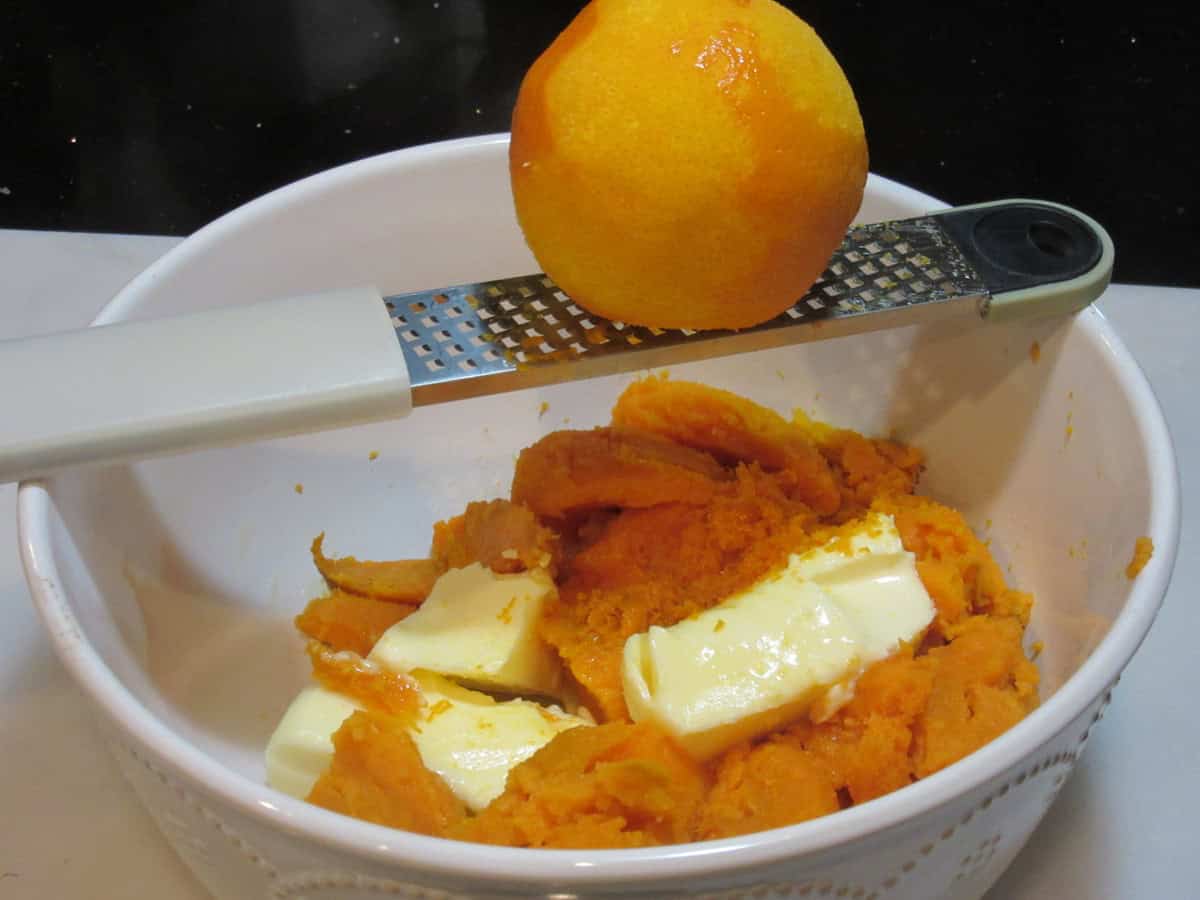 An fresh orange being zested over a bowl of cooked sweet potatoes and softened butter.