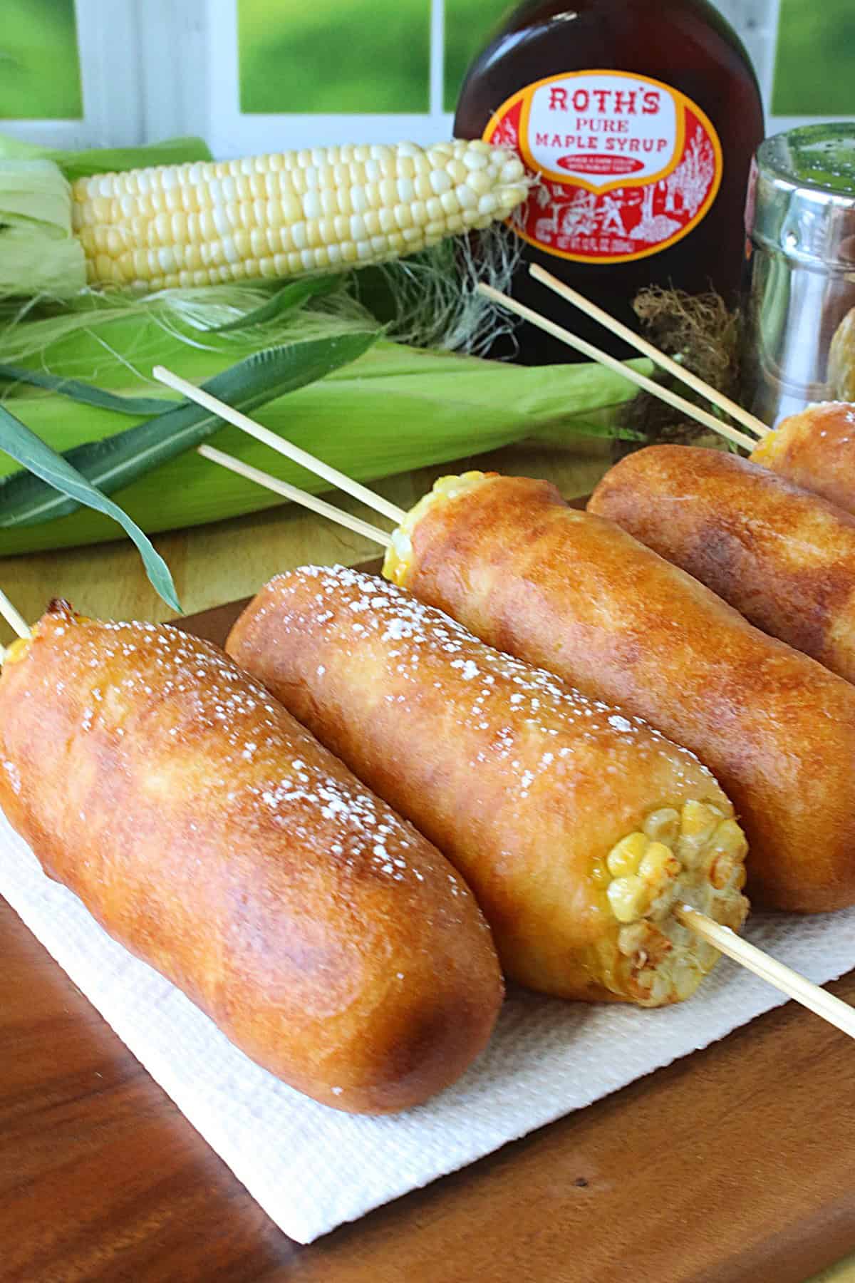 A bottle of maple syrup and a corn on the cob in the background of a lineup of corn on the cob fritters.