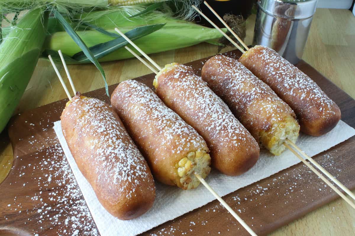 A wooden board with corn on the cob fritters topped with powdered sugar.