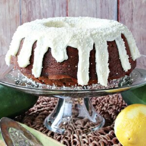 A pretty frosted Gingerbread Bundt Cake with Zucchini on a glass cake stand with a lemon a zucchini on the table.