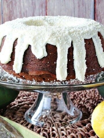 A pretty frosted Gingerbread Bundt Cake with Zucchini on a glass cake stand with a lemon a zucchini on the table.