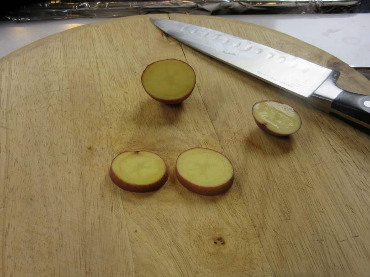 Slices of baby red potatoes on a wooden cutting board along with a knife.