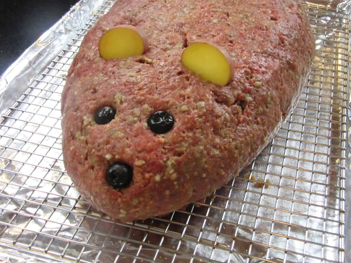 An unbaked rat loaf meatloaf with potato slice ears and olive eyes and nose.