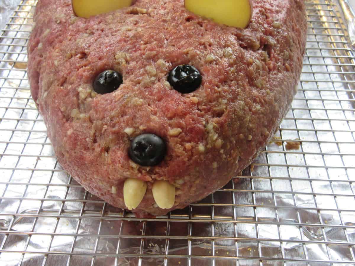 A face of an unbaked rat loaf meatloaf with olive eyes and nose along with almond teeth.