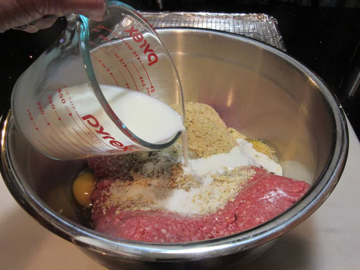 Milk being added to a meatloaf mixture in a large silver bowl.