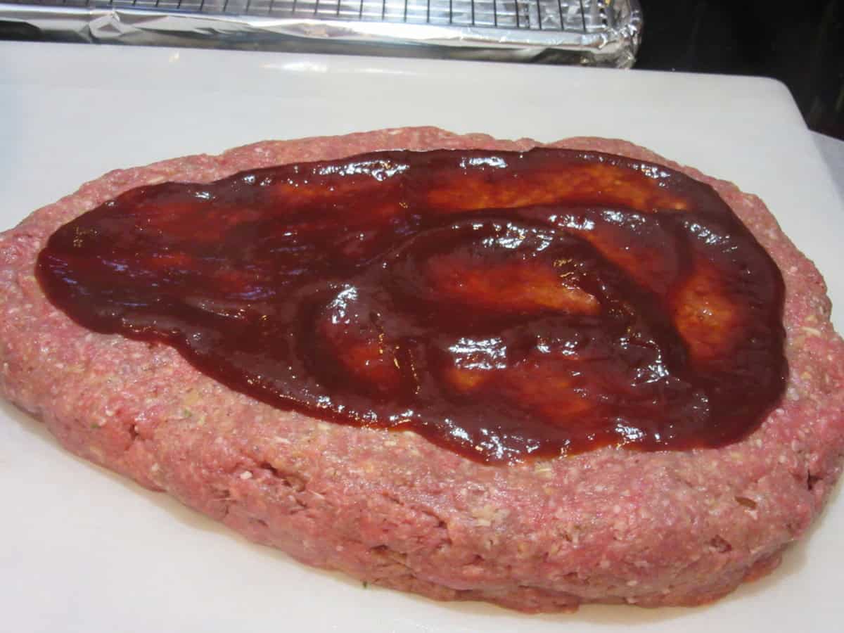 The bottom layer of an unbaked meatloaf slathered with BBQ sauce on a sheet of parchment paper.