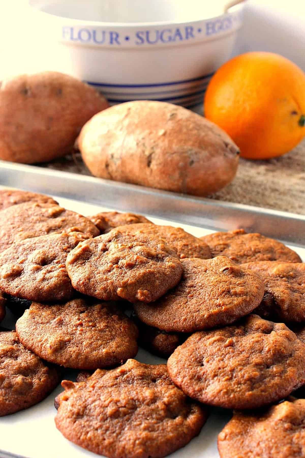 Looking for an alternative to pumpkin spice cookies this Fall? Look no further. These Amish Sweet Potato Cookies are soft and chewy with a mild spicy flavor that pairs perfectly with a cup of coffee or tea. - Kudos Kitchen by Renee Two whole sweet potatoes in the background of a baking sheet filled with Amish Sweet Potato Cookies.