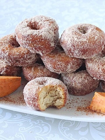 A platter of sweet potato donuts dipped in cinnamon sugar with one in the front with a bite taken out.