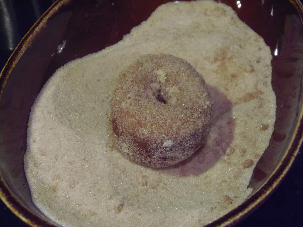 A donut just out of hot oil sitting in a bowl of cinnamon sugar for topping.