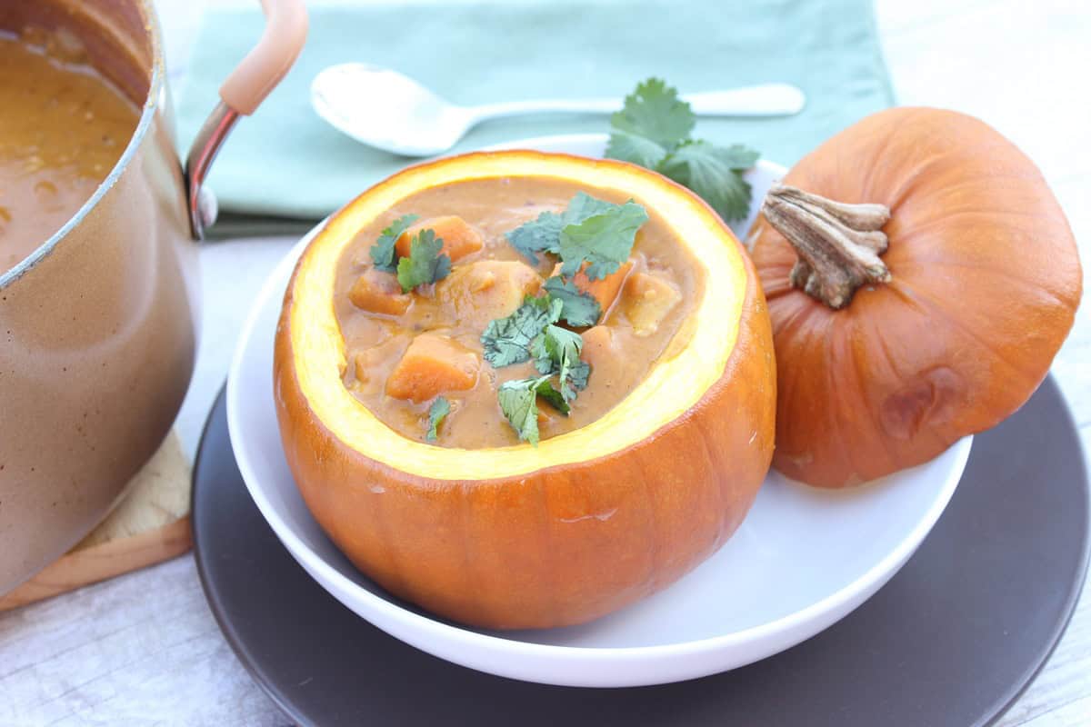 Peanut Butter, Chicken and Pumpkin Stew A small pumpkin filled with chicken pumpkin stew on a white plate with a spoon in the background.