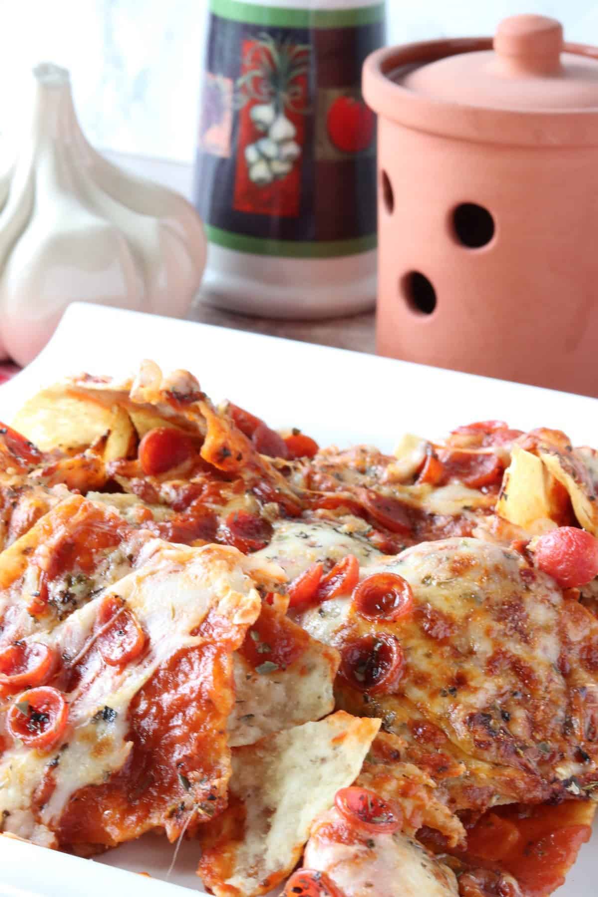 A platter of pizza nachos in the foreground with a garlic keeper in the background.