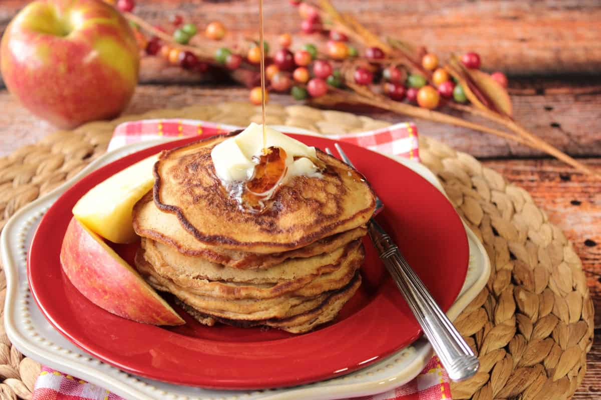 The flavors of fall apples come alive in these pancakes making them delicious for breakfast, or for a treat on those breakfast-for-dinner nights. - Kudos Kitchen by Renee A red plate with a stack of apple cider pancakes and a drizzle of maple syrup from above.