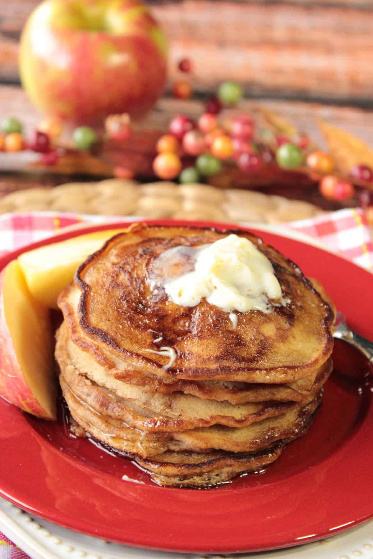 The flavors of fall apples come alive in these pancakes making them delicious for breakfast, or for a treat on those breakfast-for-dinner nights. - Kudos Kitchen by Renee An apple and fall berries in the background of a plate of apple cider pancakes with butter.