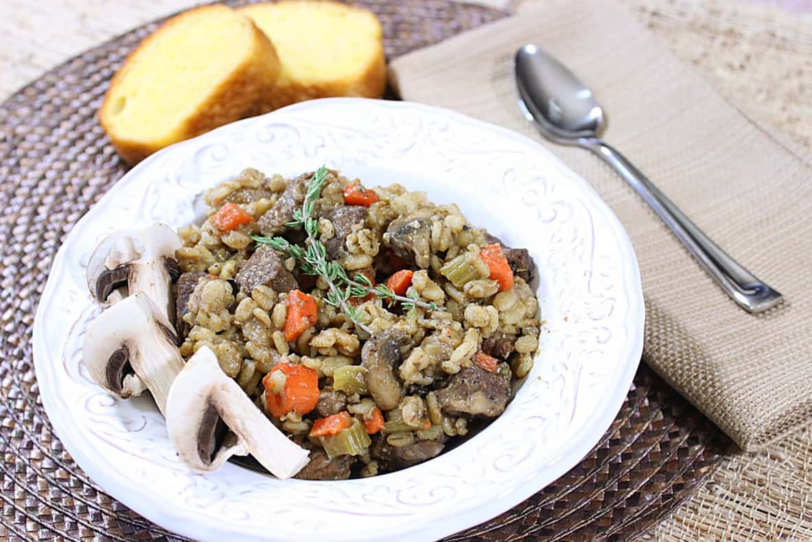 A spoon on a tan napkin next to a bowl of beef and barley stew with carrots.
