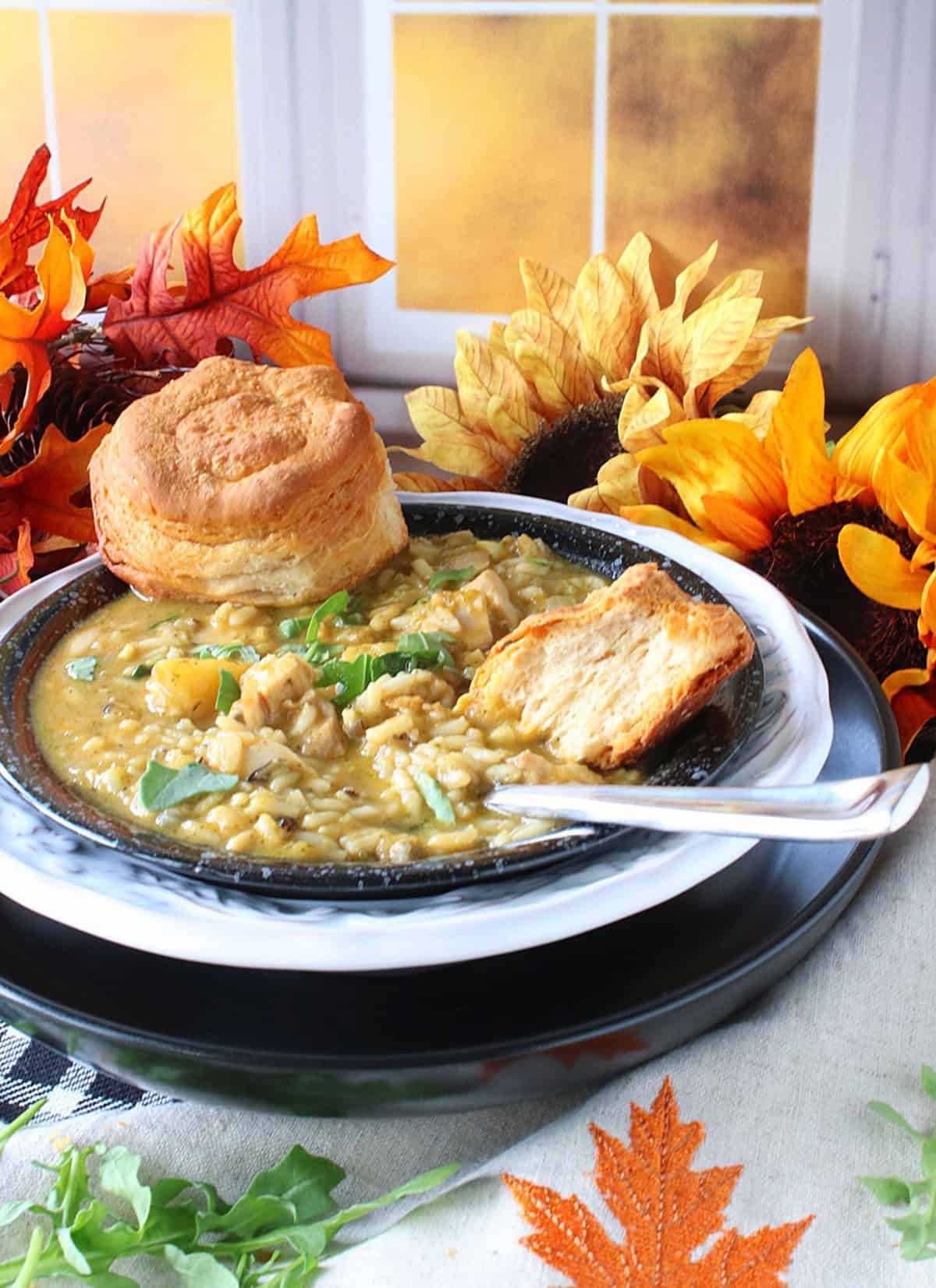 A fall setting around a bowl of creamy chicken and wild rice soup topped with arugula and biscuits.