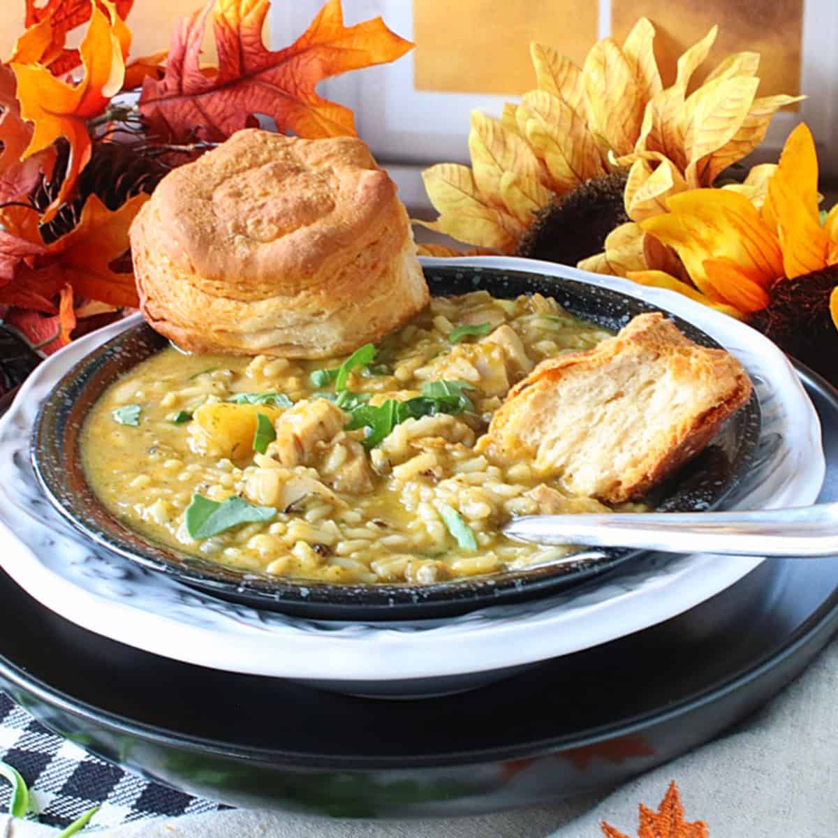 A black bowl with chicken and wild rice soup and a spoon in front of a fall setting with leaves and sunflowers.