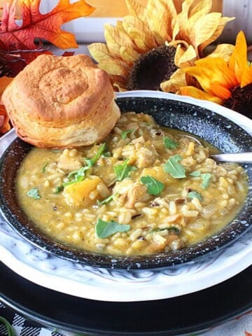 A creamy bowl of chicken and wild rice soup topped with arugula, a biscuit and a spoon inside the bowl.