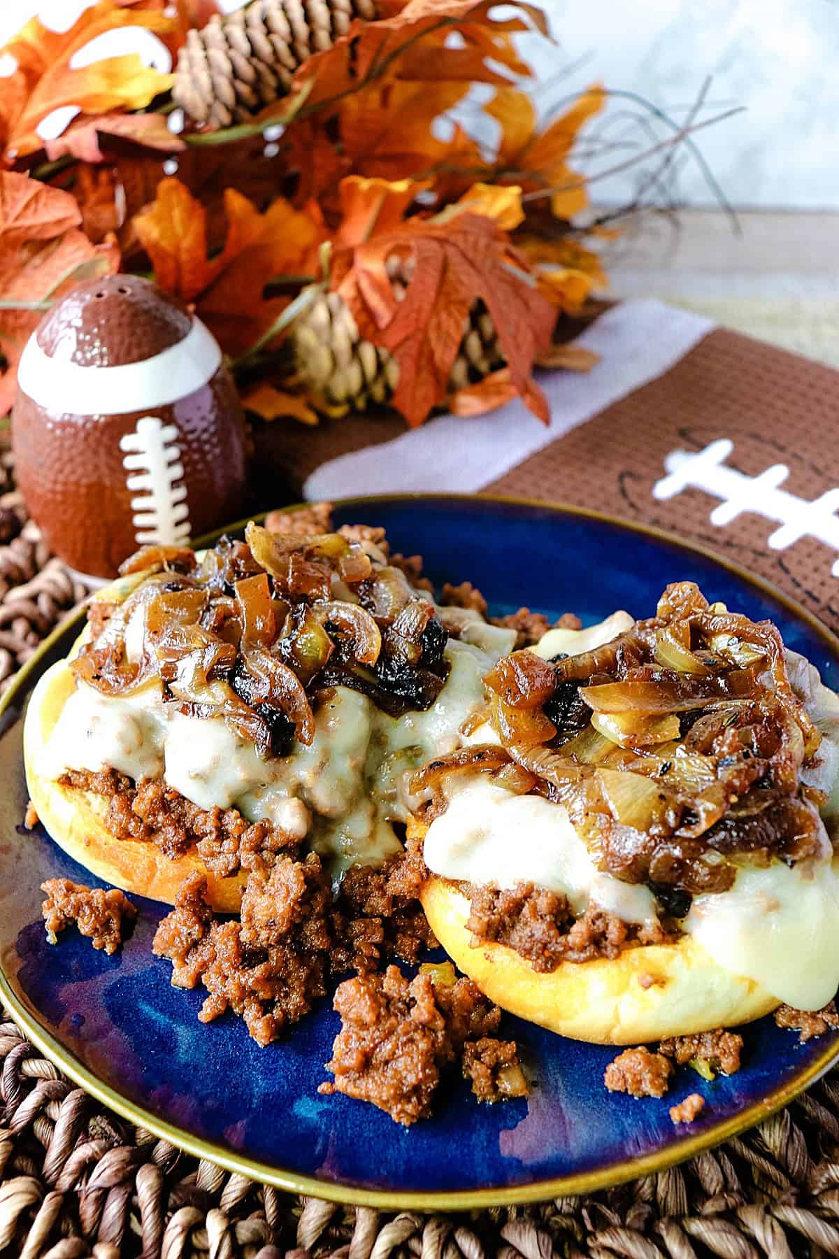 A blue plate with an open face French onion sloppy joe sandwich and a fall and football theme in the background.