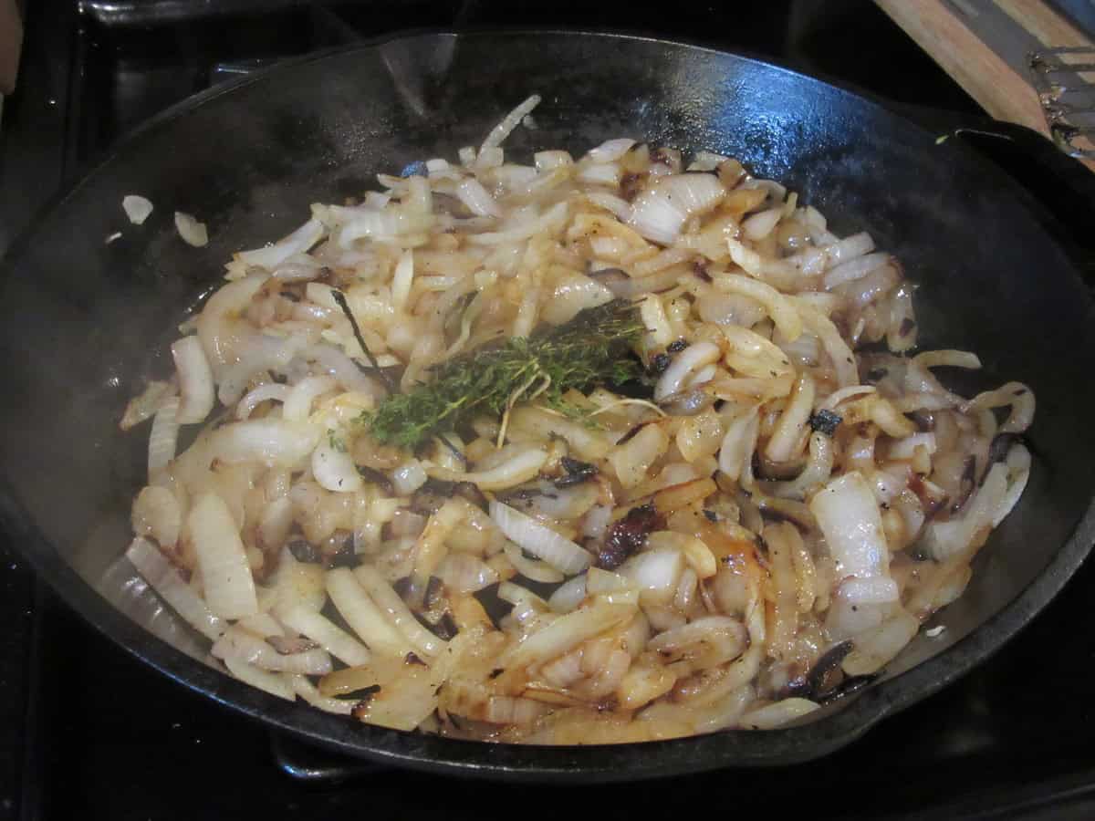 Cooking caramelized onions with a thyme sprig bouquet in a cast iron skillet on the stovetop.