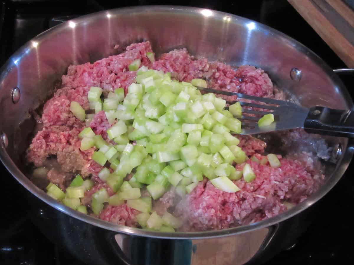 Raw ground beef and diced celery in a large skillet with a slotted spatula.